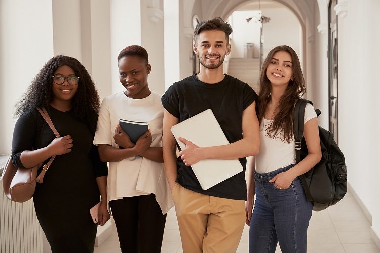 Front view of four happy students with backpacks going to lecture, standing on corridor and smiling at camera. Caucasian and African friends studying together at university, resting on break.